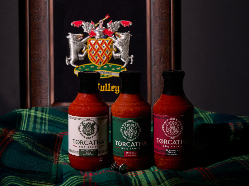 An image of three Torcatha BBQ sauces, one with a white label, one with a green label, and one with a red label, sitting on Scottish plaid fabric with the McCully family crest in the background.