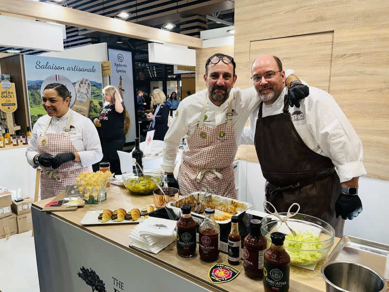 Three chefs in a kitchen setting with brisket sandwiches, bottles of barbecue sauce, and several mixing bowls on a counter.