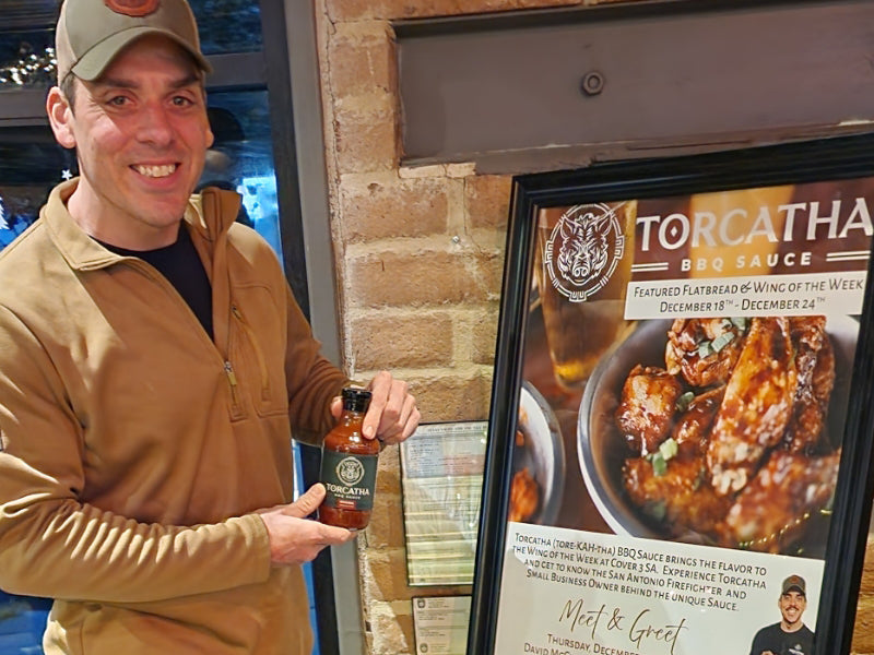 Man holding a bottle of Torcatha BBQ Sauce, the best barbecue sauce for wings, in front of a promotional poster for "The Wing of the Week."