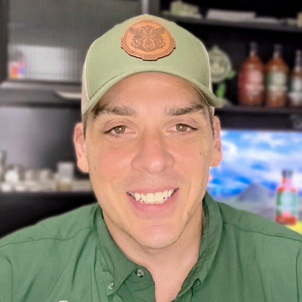 Man wearing a green cap with an illustration of a boar, representing Torcatha BBQ Sauce, and green shirt in an indoor setting with bottles of the best barbecue sauce in the background.