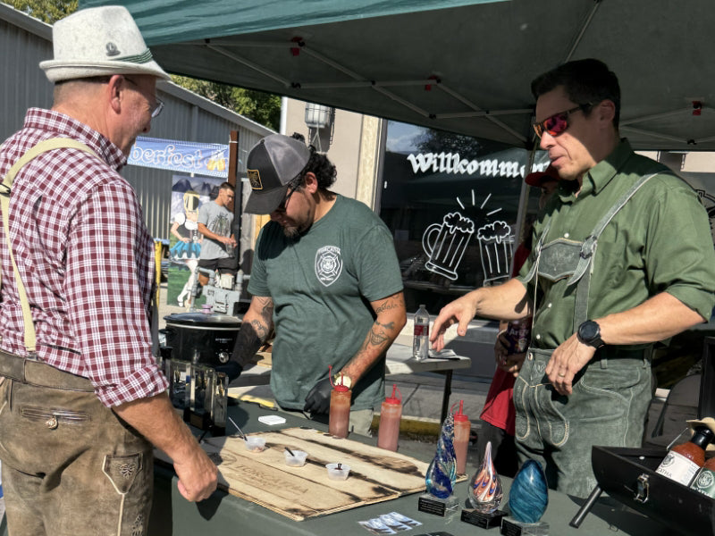 Group of three men wearing lederhosen and sampling BBQ sauces at a food festival booth.