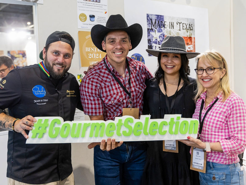Four people, two wearing cowboy hats and one in a chef's uniform, holding a "#GourmetSelection" sign at an event with "Made in Texas" promotional materials in the background.