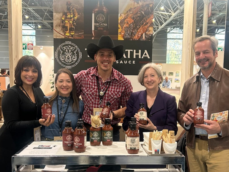 Group of people wearing Western-style clothing holding Torcatha BBQ Sauce bottles and promotional materials at a food show booth.