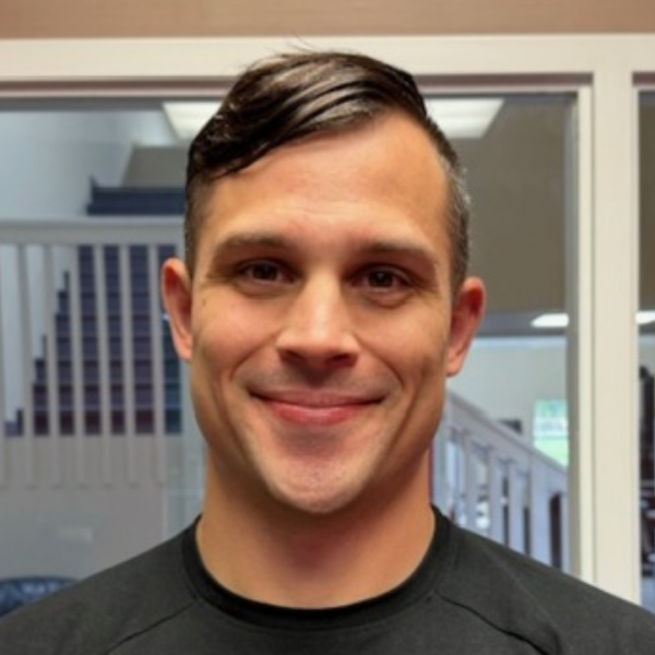 A man wearing a black T-shirt, smiling while standing in front of a staircase with a white railing.