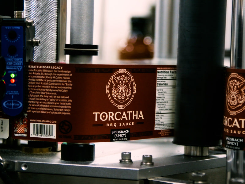 A labeling machine with a red label featuring a boar's head, stretched across the rollers, reading: "Torcatha BBQ Sauce Spìosrach (Spicy)."