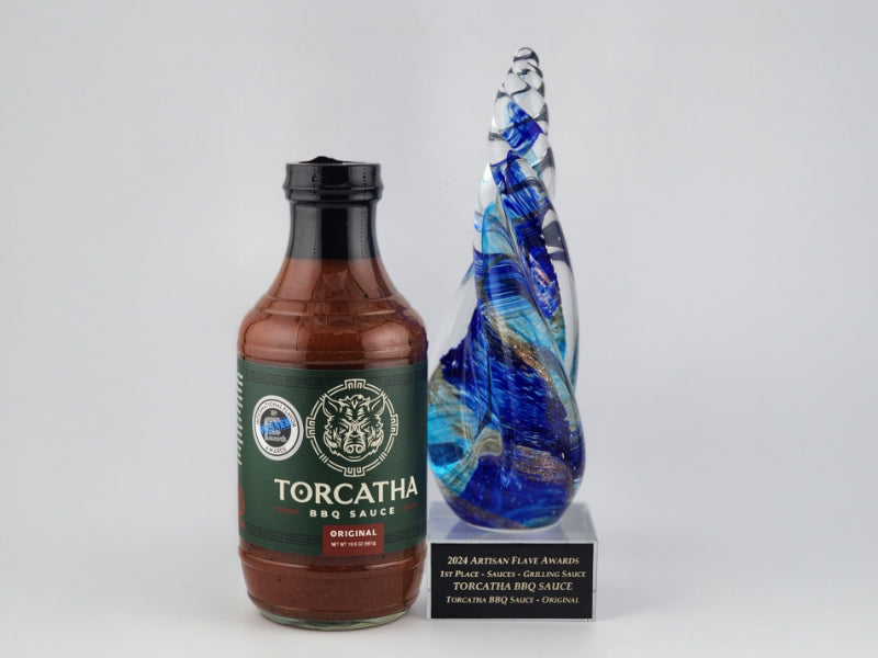 A bottle Original Torcatha BBQ Sauce next to a blue decorative glass trophy that reads, "2024 Artisan Flave Awards, 1st Place Sauces, Grilling Sauce, Torcatha BBQ Sauce."