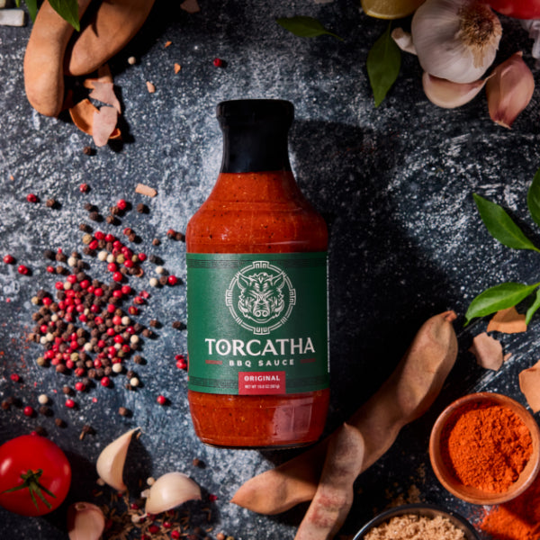 An Original flavor Torcatha BBQ Sauce bottle surrounded by red, brown, and orange spices, along with a garlic bulb, tomatoes, leaves, and other ingredients on a dark surface.