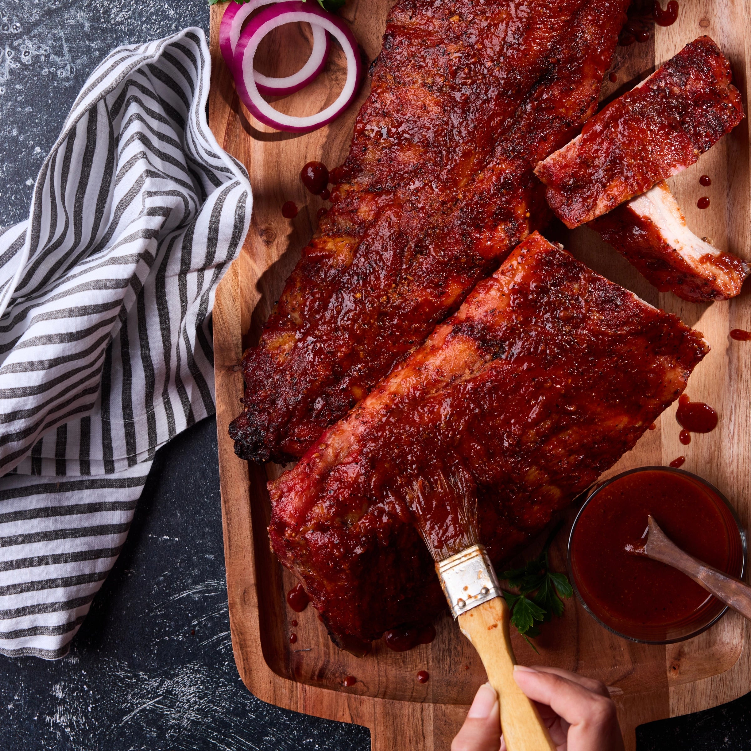 Barbecue ribs on a wooden cutting board with a brush and BBQ sauce, next to a blue and white striped towel.