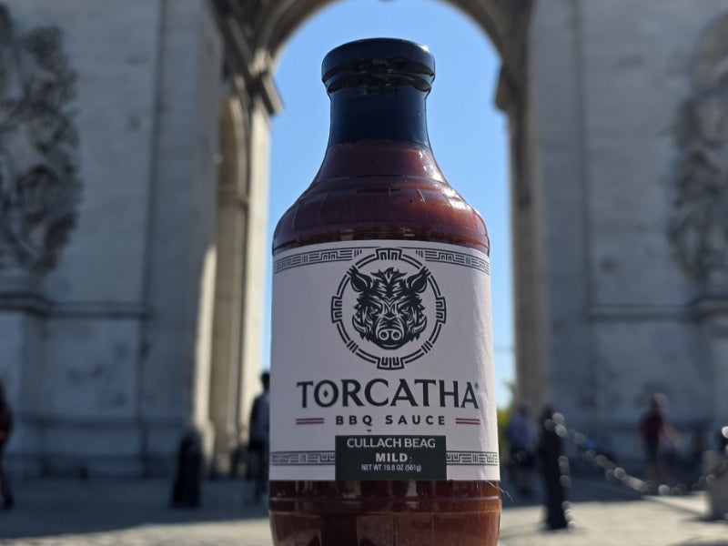 Bottle of Torcatha Cullah Beag Mild BBQ Sauce held in front of the Arc de Triomphe, in Paris, with people in the background.