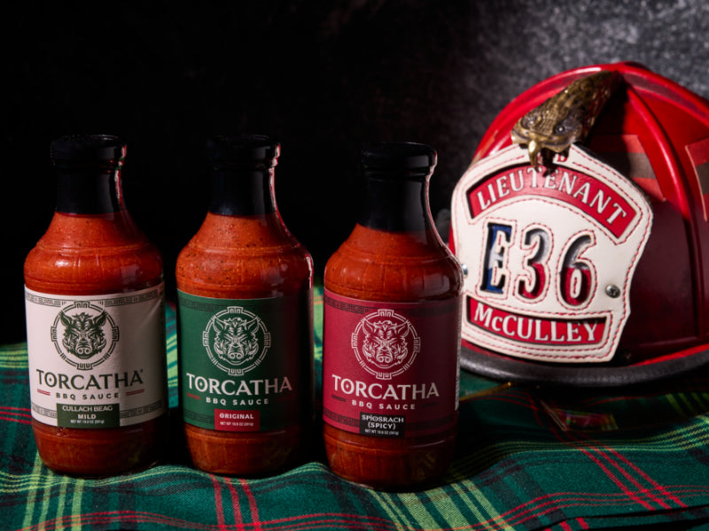 Three bottles of BBQ sauce, one with a white label, one with a green label, and one with a red label sitting on Scottish plaid fabric next to a fire helmet with text that reads, "Lieutenant McCully E36."