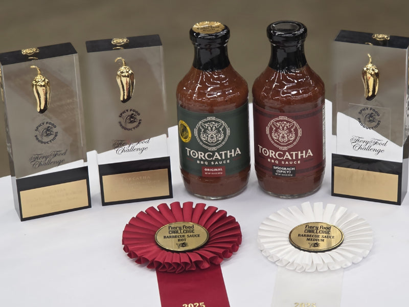 Two bottles of Torcatha barbecue sauce with three glass trophies and two award ribbons on a white table.