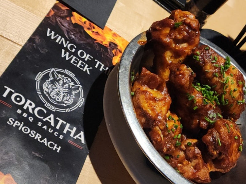 A stainless steel bowl filled with barbecue chicken wings garnished with scallions, sitting on an oak-colored table next to a menu that reads, "Wing of the Week: Torcatha BBQ Sauce Spìosrach."