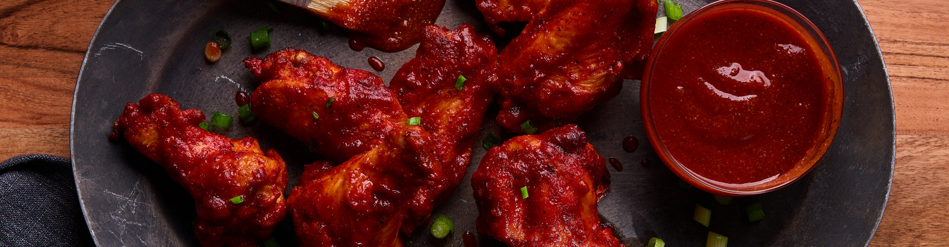 A black plate on a wooden table with BBQ chicken wings garnished with scallions, next to a small bowl of BBQ sauce.