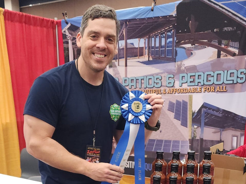 A man smiling and holding a blue ribbon in front of a "Patio & Pergolas" display, wearing a navy blue shirt and a Zest Fest lanyard.