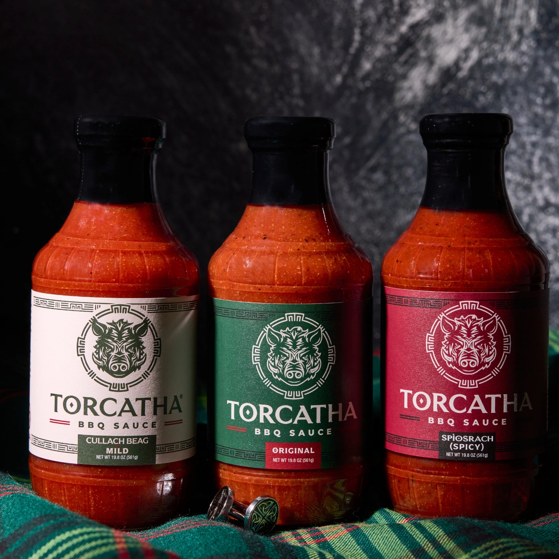 Three bottles of Torcatha BBQ Sauce on a plaid surface with a dark background, featuring: Cullach Beag (Mild), Original, and Spìosrach (Spicy).