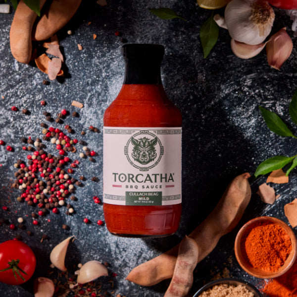 A Torcatha BBQ Sauce bottle, Cullach Beag (Mild flavor), surrounded by red, brown, and orange spices, along with a garlic bulb, tomatoes, leaves, and other ingredients on a dark surface.