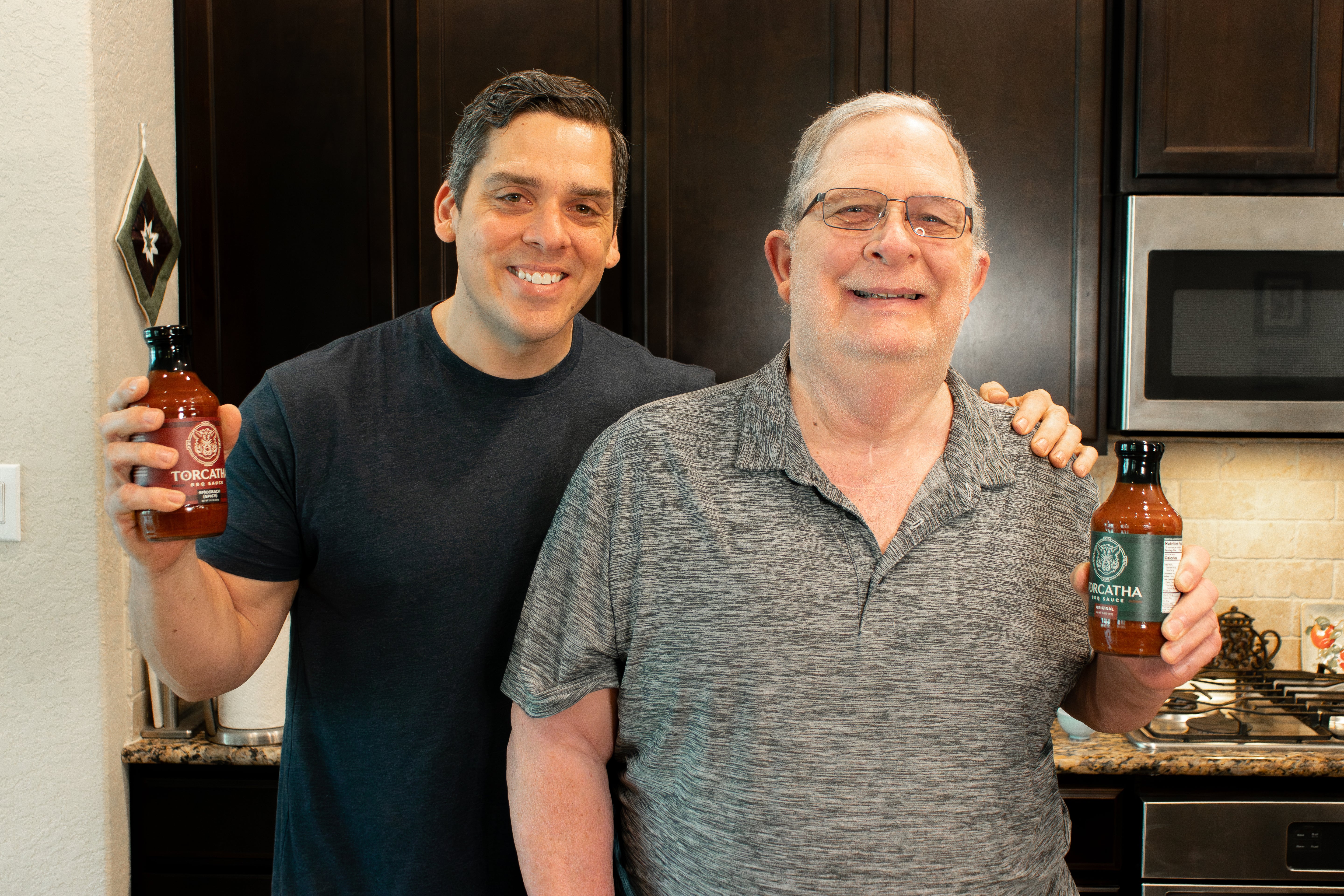 An older man and a younger man standing side by side in a kitchen, each holding a bottle of the best barbecue sauce for barbecue ribs and smiling at the camera.