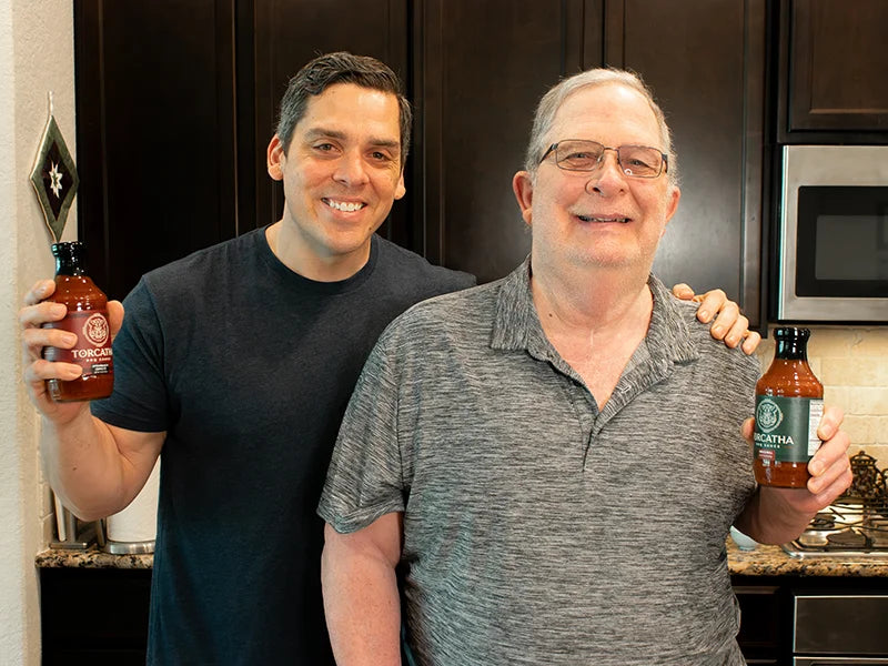 An older man and a younger man standing side by side in a kitchen, each holding a bottle of the best barbecue sauce for pulled pork and smiling at the camera.
