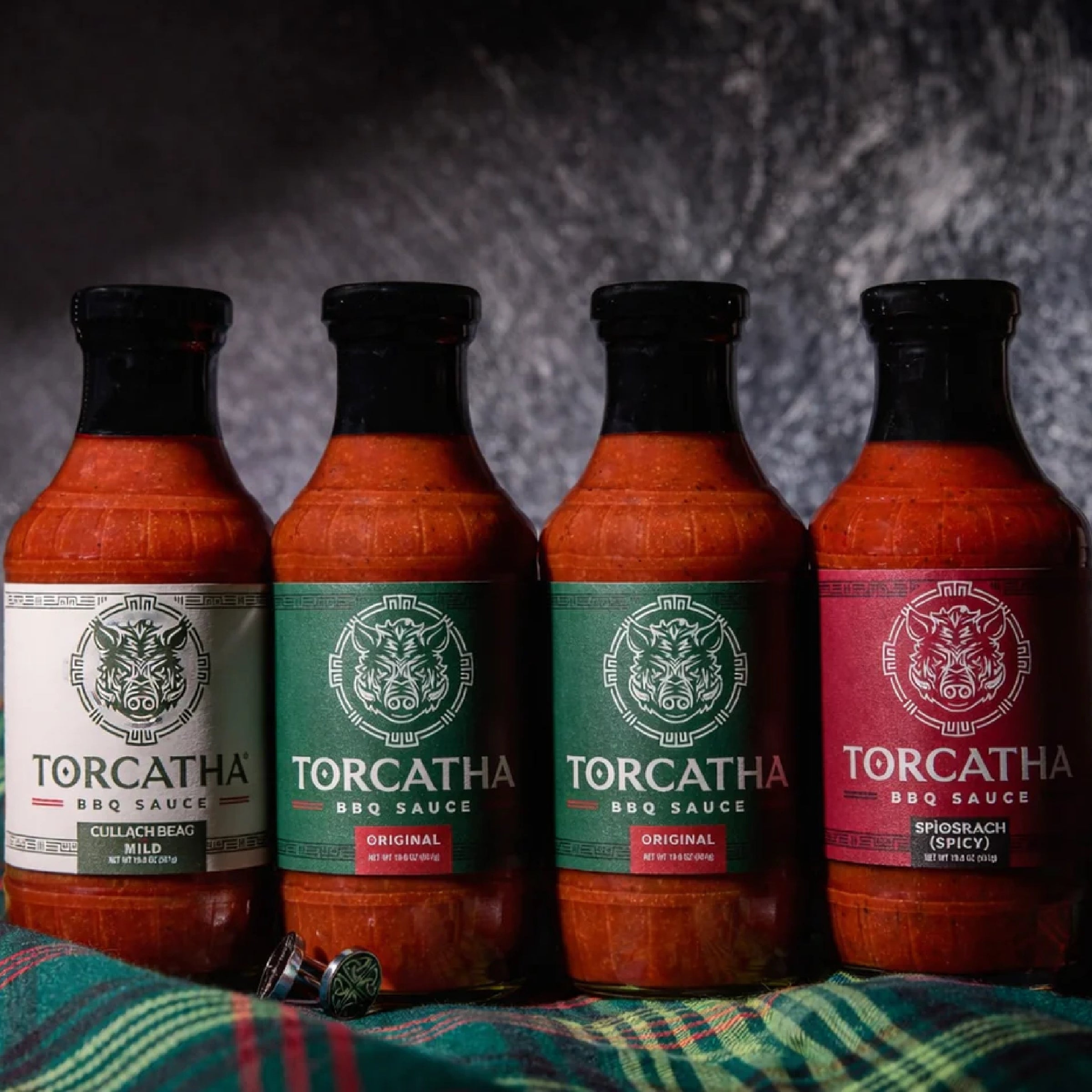 An image of four Torcatha BBQ sauces, one with a white label, two with green labels, and one with a red label, sitting on Scottish plaid fabric.