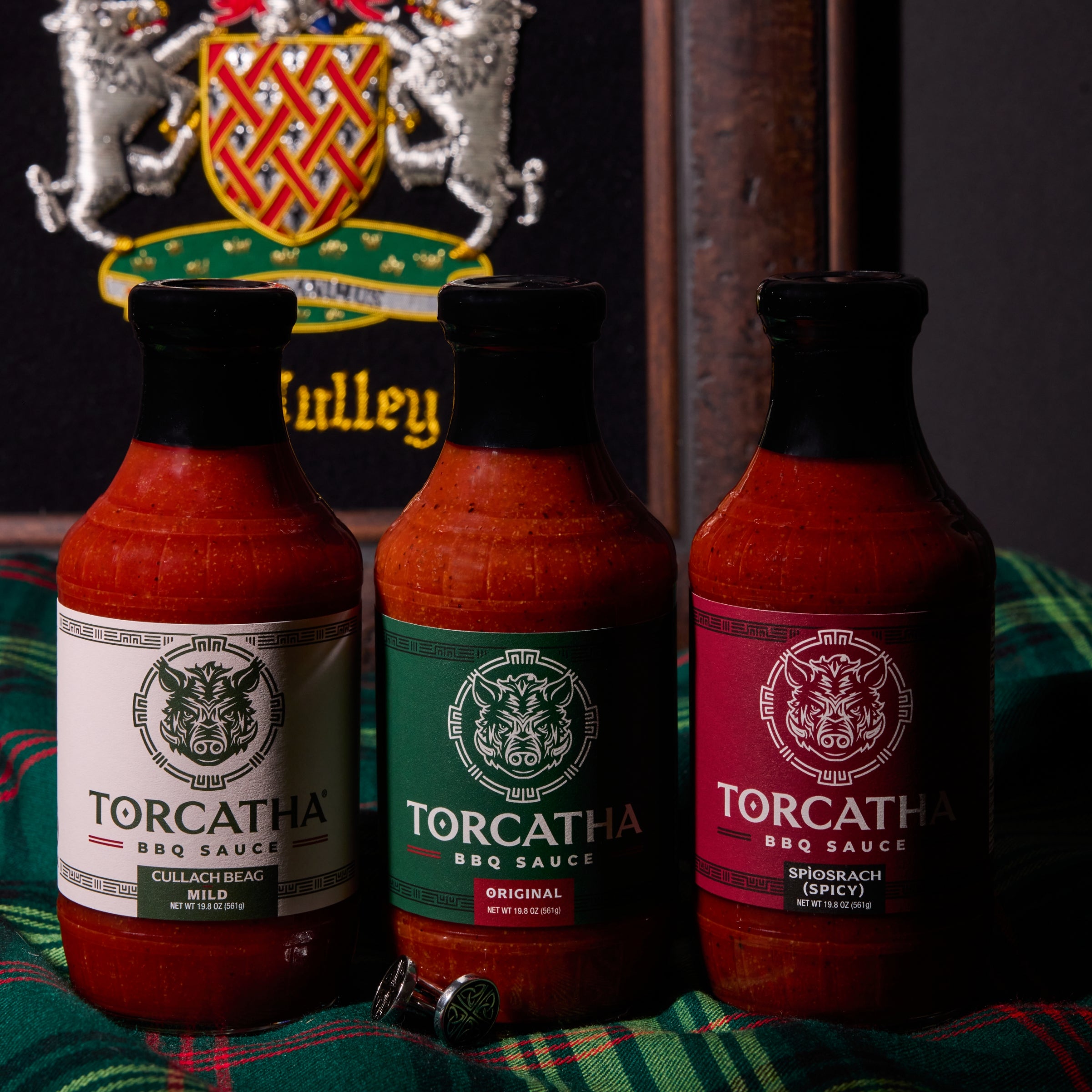 An image of three Torcatha BBQ sauces, one with a white label, one with a green label, and one with a red label, sitting on Scottish plaid fabric with the McCully family crest in the background.