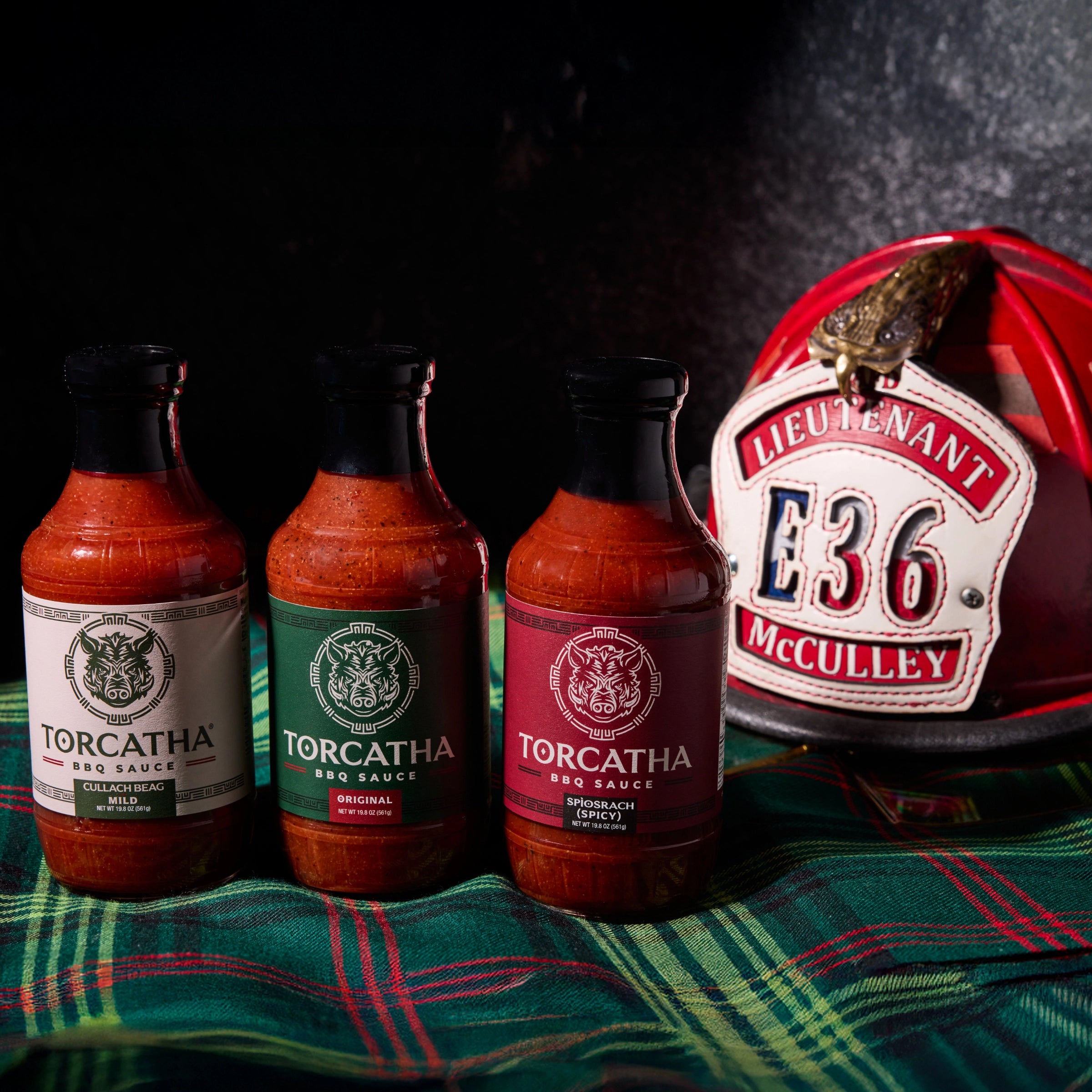 Three bottles of BBQ sauce, one with a white label, one with a green label, and one with a red label sitting on Scottish plaid fabric next to a fire helmet with text that reads, "Lieutenant McCully E36."