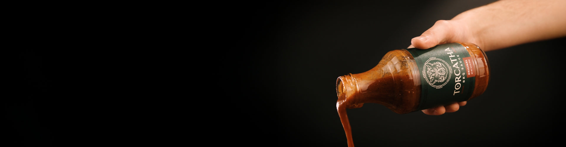  A hand pouring a bottle of Torcatha Original barbecue sauce with a dark background.