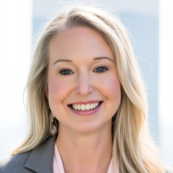 Woman with blonde hair and blue eyes, wearing a grey blazer and drop earrings, smiling at the camera against a light blue background.