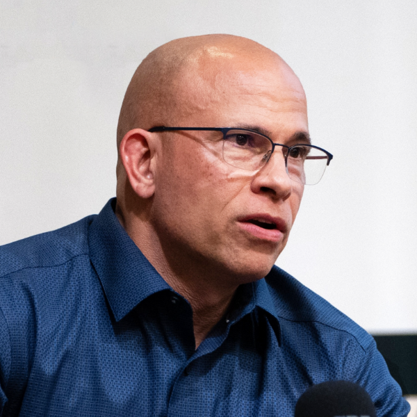 A bald, middle-aged man wearing glasses and a blue shirt speaking into a microphone against a white background.