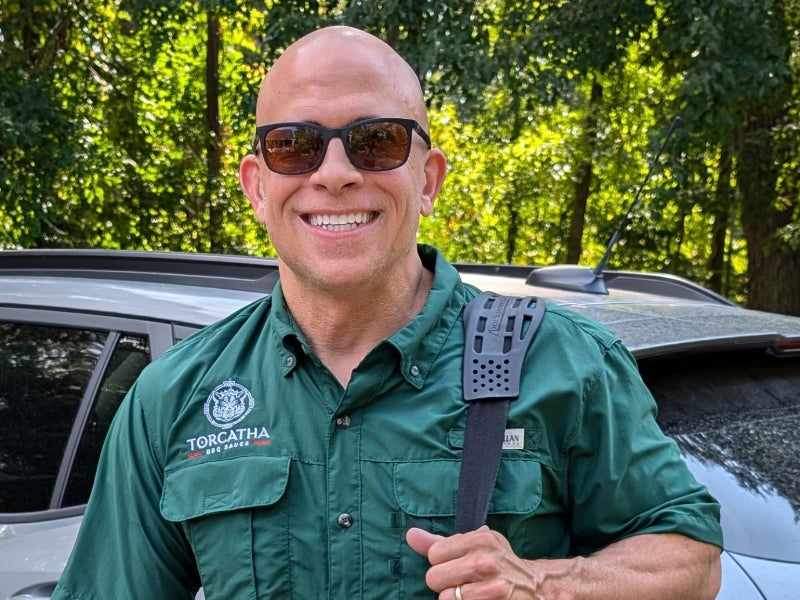 Man in green shirt with 'Torcatha' logo, smiling and wearing sunglasses  holding a bag, standing in front of a vehicle with trees in the background.