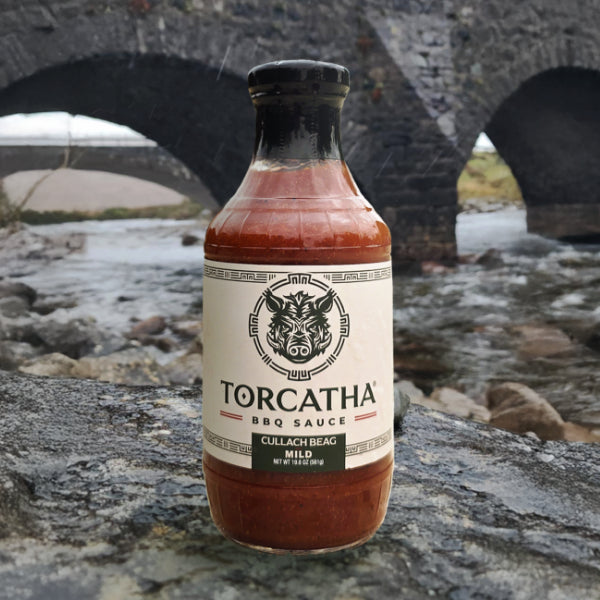 A bottle of Torcatha Cullach Beag Mild BBQ Sauce sitting on a rock, with a river and a stone bridge in the background. 