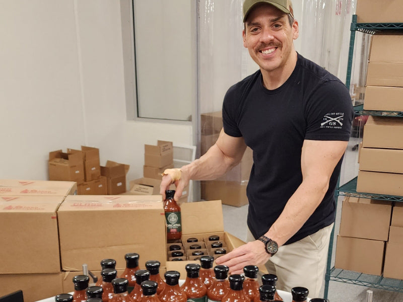 Man in a warehouse packing boxes of barbecue sauce, wearing a black t-shirt and green cap, and smiling at the camera.