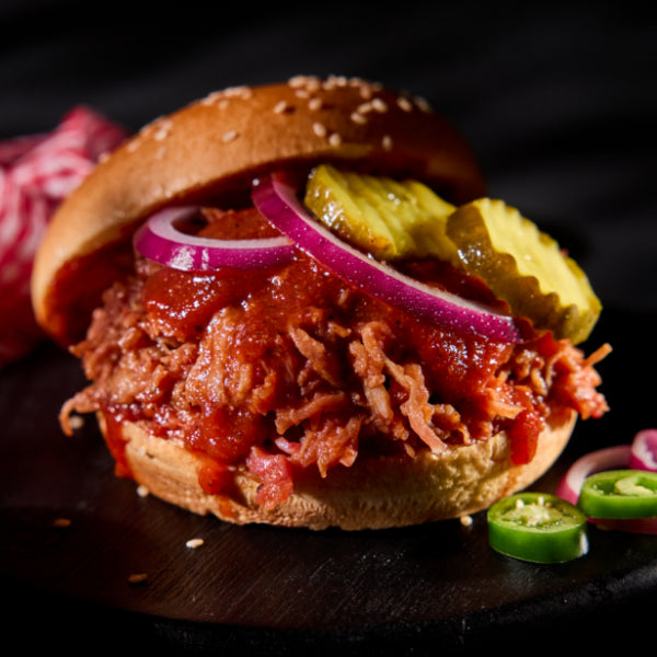 Pulled pork sandwich with pickles and onions on a sesame seed bun, placed next to jalapeños on a dark wood table.
