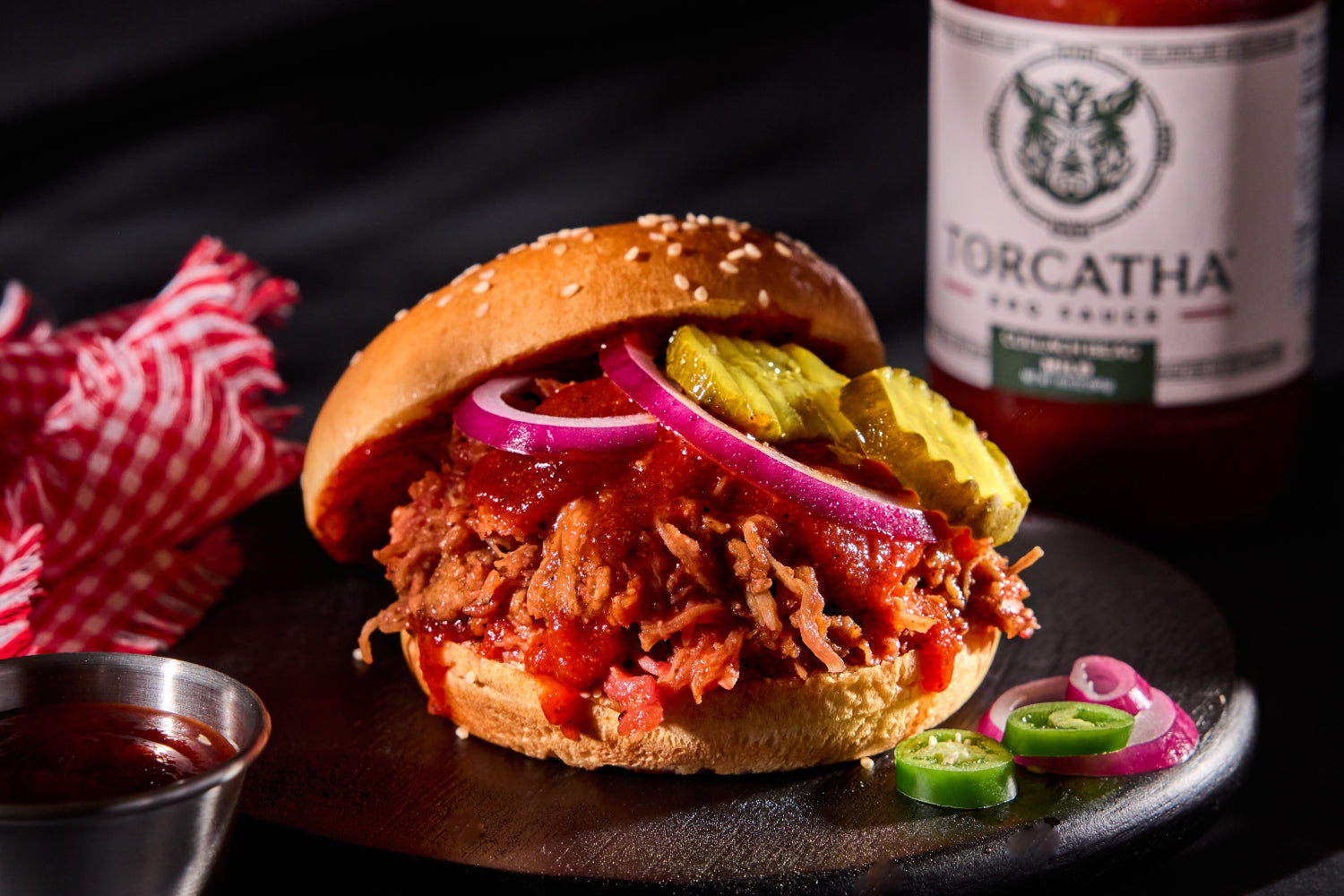 Pulled pork sandwich with pickles and onions on a bun, accompanied by a bottle of Torcatha barbecue sauce, the best BBQ sauce for pulled pork.