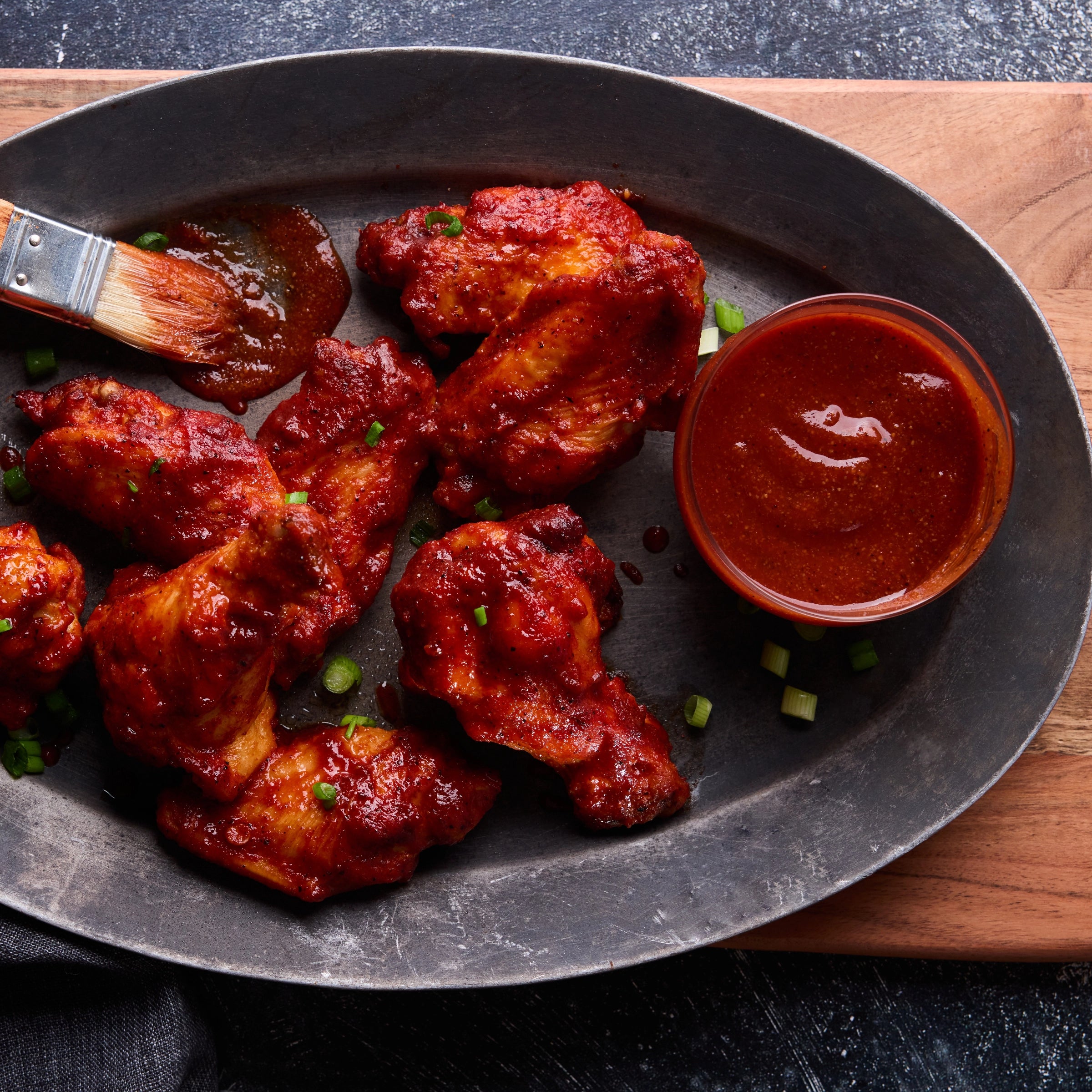 Chicken wings on a black, oval plate, garnished with scallions, served with a side of Torcatha barbecue sauce on a wooden cutting board, the best BBQ sauce for wings.