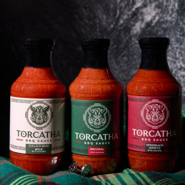 Three bottles of Torcatha BBQ Sauce on a plaid surface with a dark background, featuring the best Texas barbecue sauces: Cullach Beag (Mild), Original, and Spìosrach (Spicy).