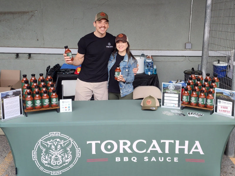 A man and a woman wearing black t-shirts and matching trucker caps stand behind a Torcatha BBQ Sauce display table with bottles and promotional materials.