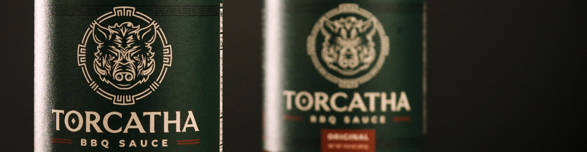 Close-up of two Torcatha BBQ Sauce bottles, with one in sharp focus and the other slightly blurred, set against a dark background.