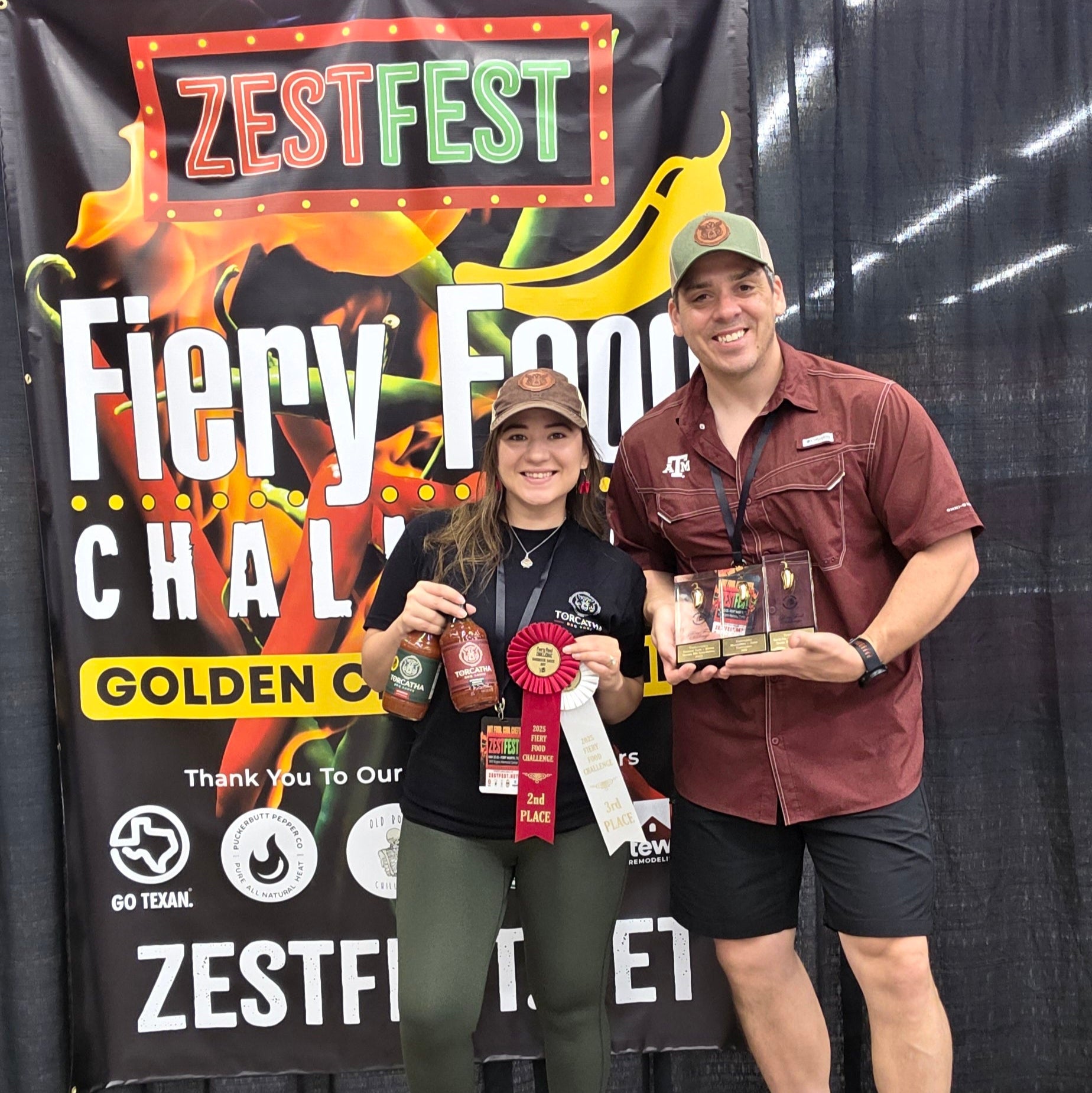 A man and a woman holding trophies, ribbons, and bottles of barbecue sauce in front of a Zest Fest Fiery Food Challenge banner. 